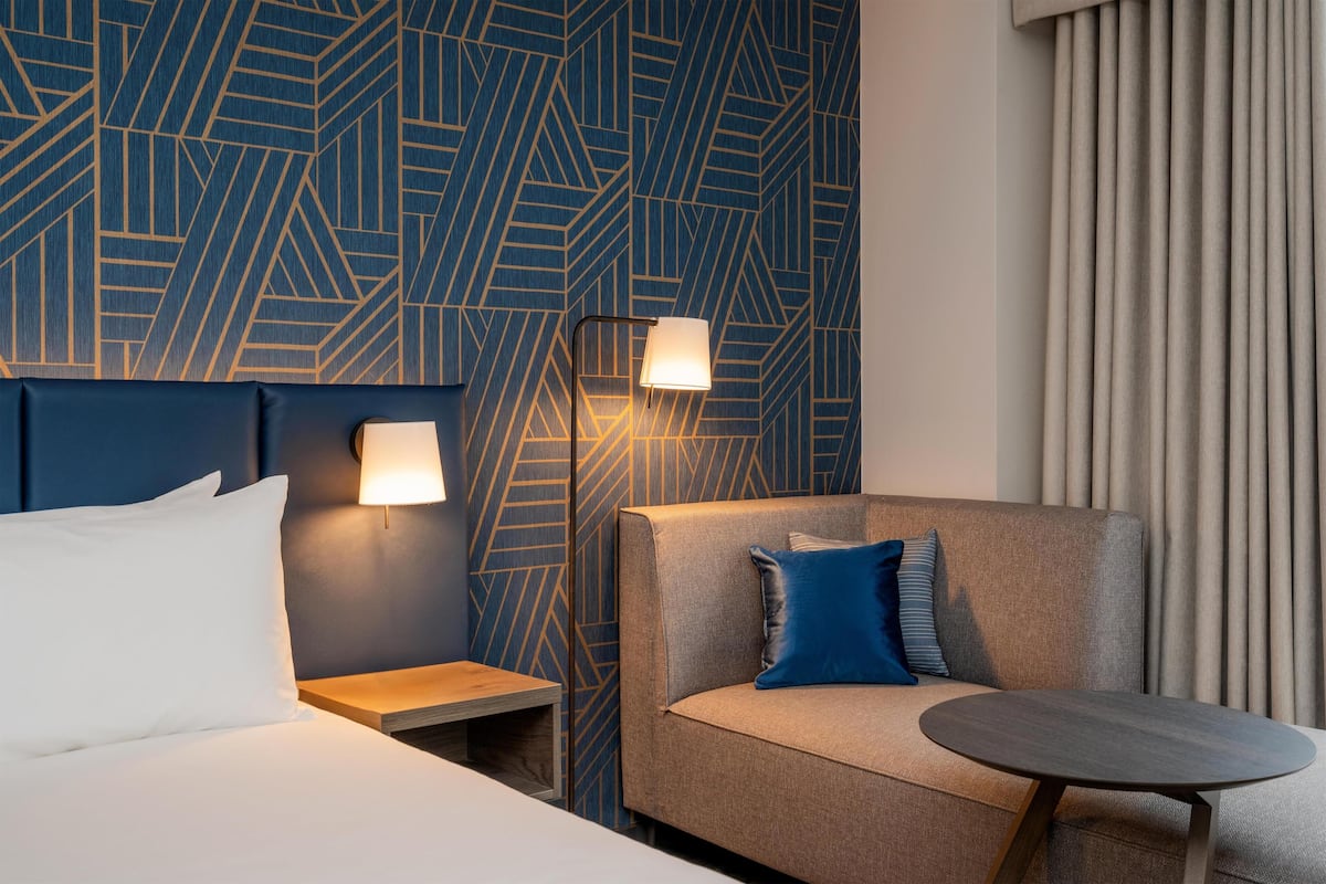 A hotel room with a bed, a sofa, a small table, and two wall-mounted lamps.