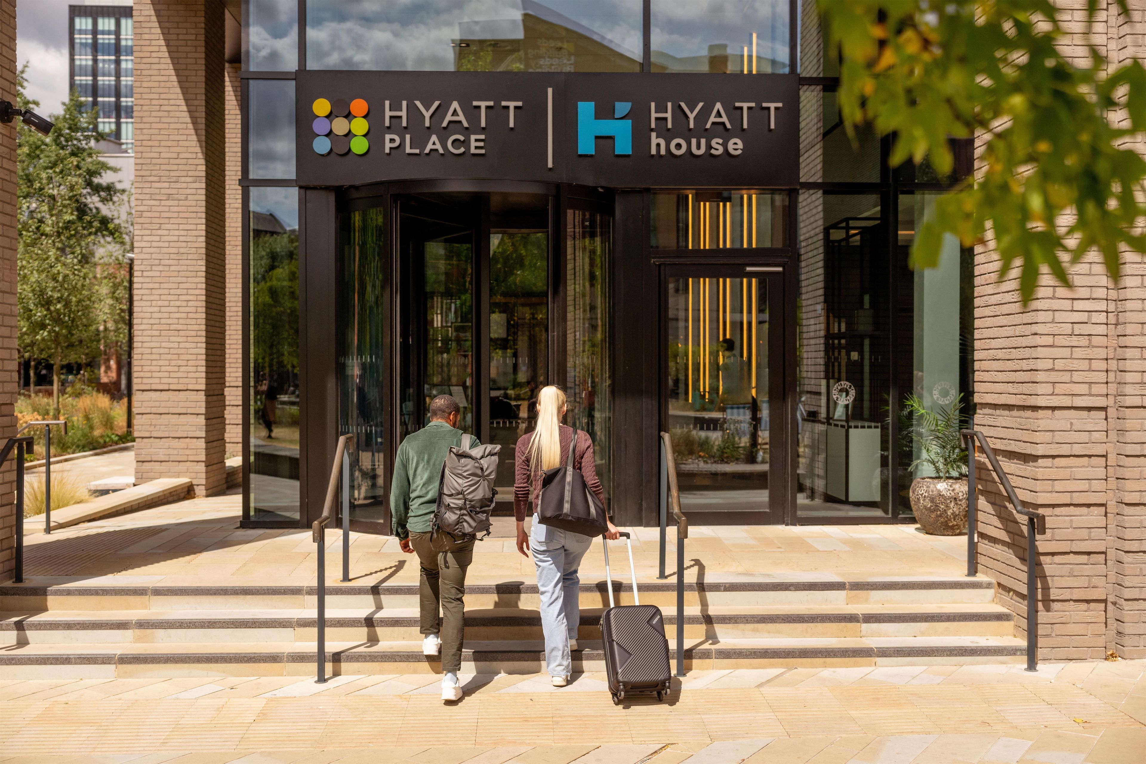 A hotel entrance with a glass facade, a sign indicating it's a Hyatt Place, and a person with a suitcase.