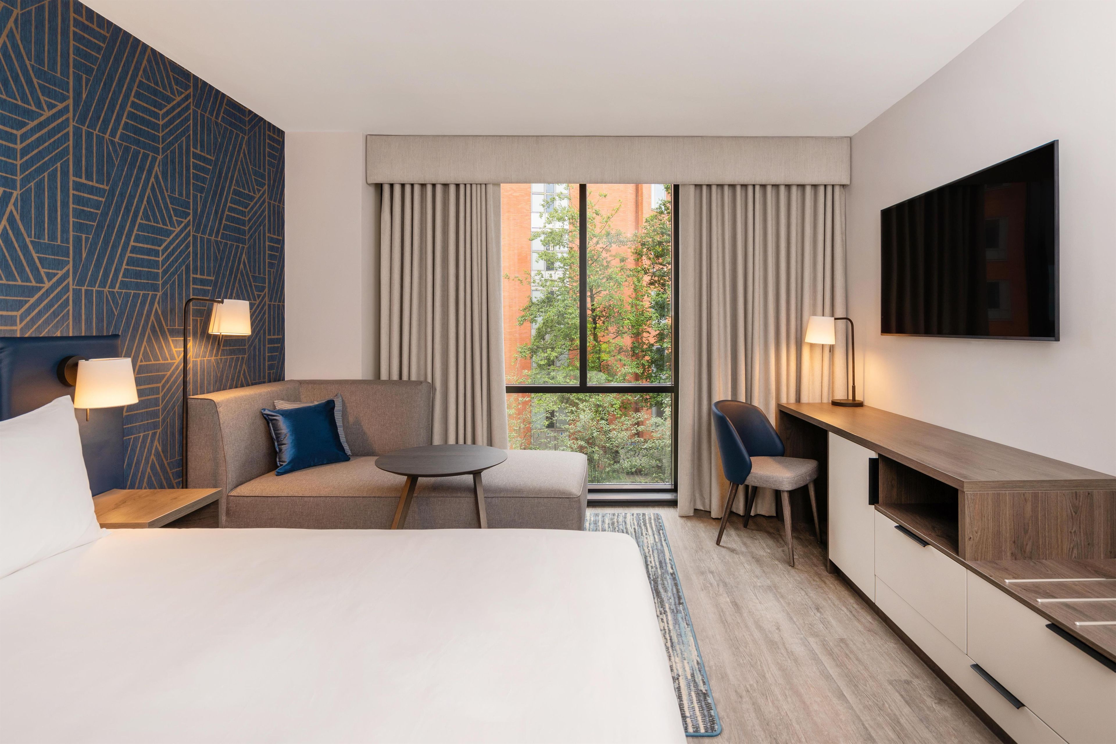 A hotel room with a bed, a sofa, a desk, and a TV. There is a window with curtains, a lamp, and a view of trees and buildings.