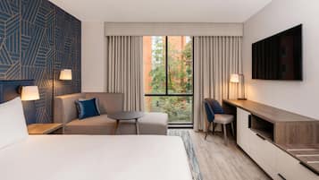 A hotel room with a bed, a sofa, a desk, and a TV. There is a window with curtains, a lamp, and a view of trees and buildings.