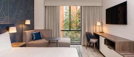 A hotel room with a bed, a sofa, a desk, and a TV. There is a window with curtains, a lamp, and a view of trees and buildings.