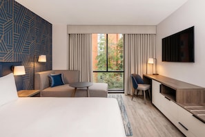 A hotel room with a bed, a sofa, a desk, and a TV. There is a window with curtains, a lamp, and a view of trees and buildings.