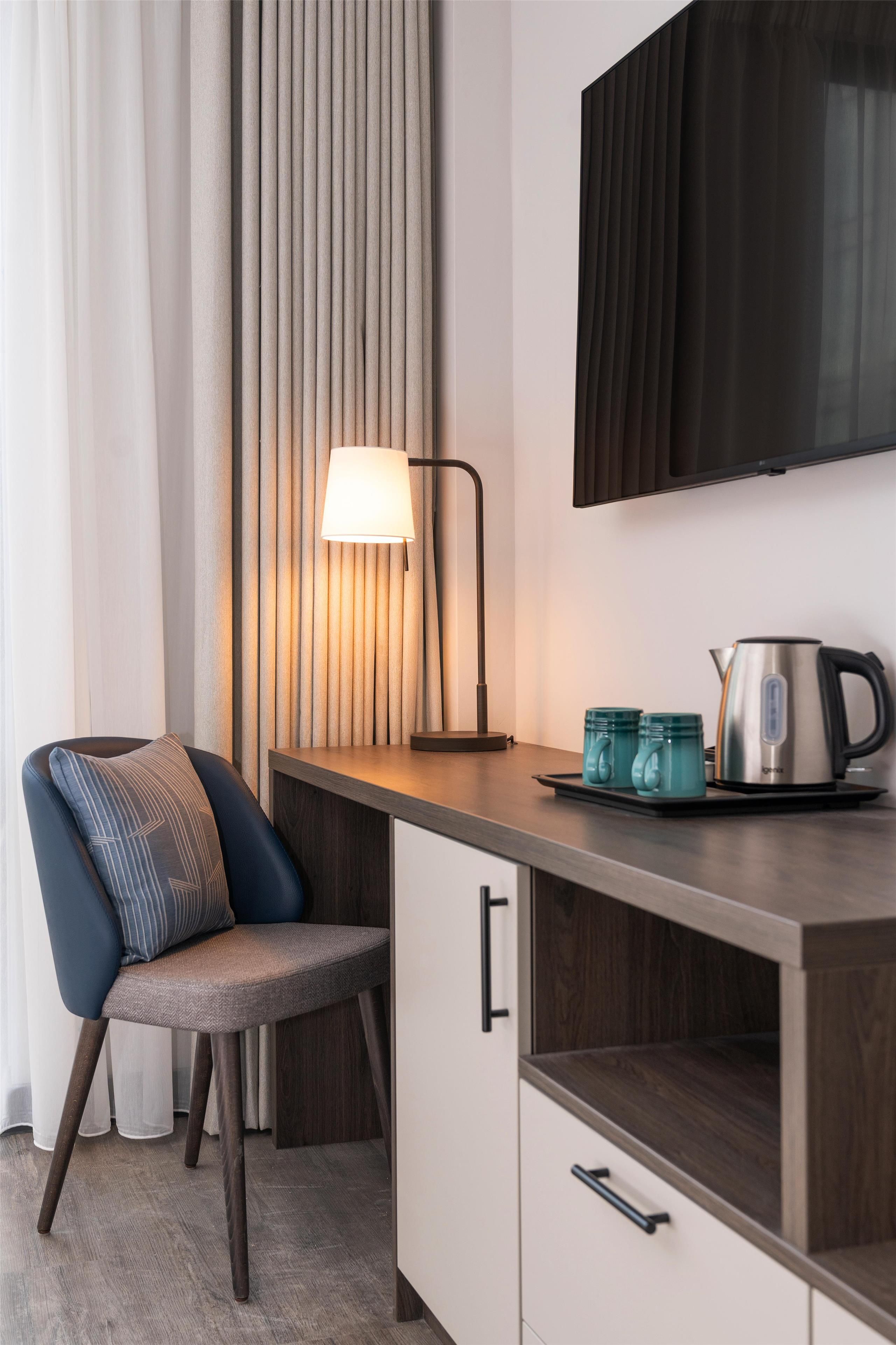 A modern kitchenette with a chair, a lamp, a kettle, and a flat-screen TV mounted on the wall.