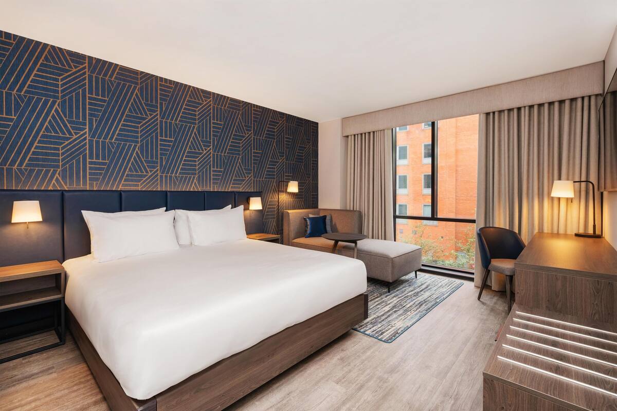 A modern hotel room with a large bed, a desk, a chair, and a sofa. There is a patterned wall behind the bed and a window with curtains.