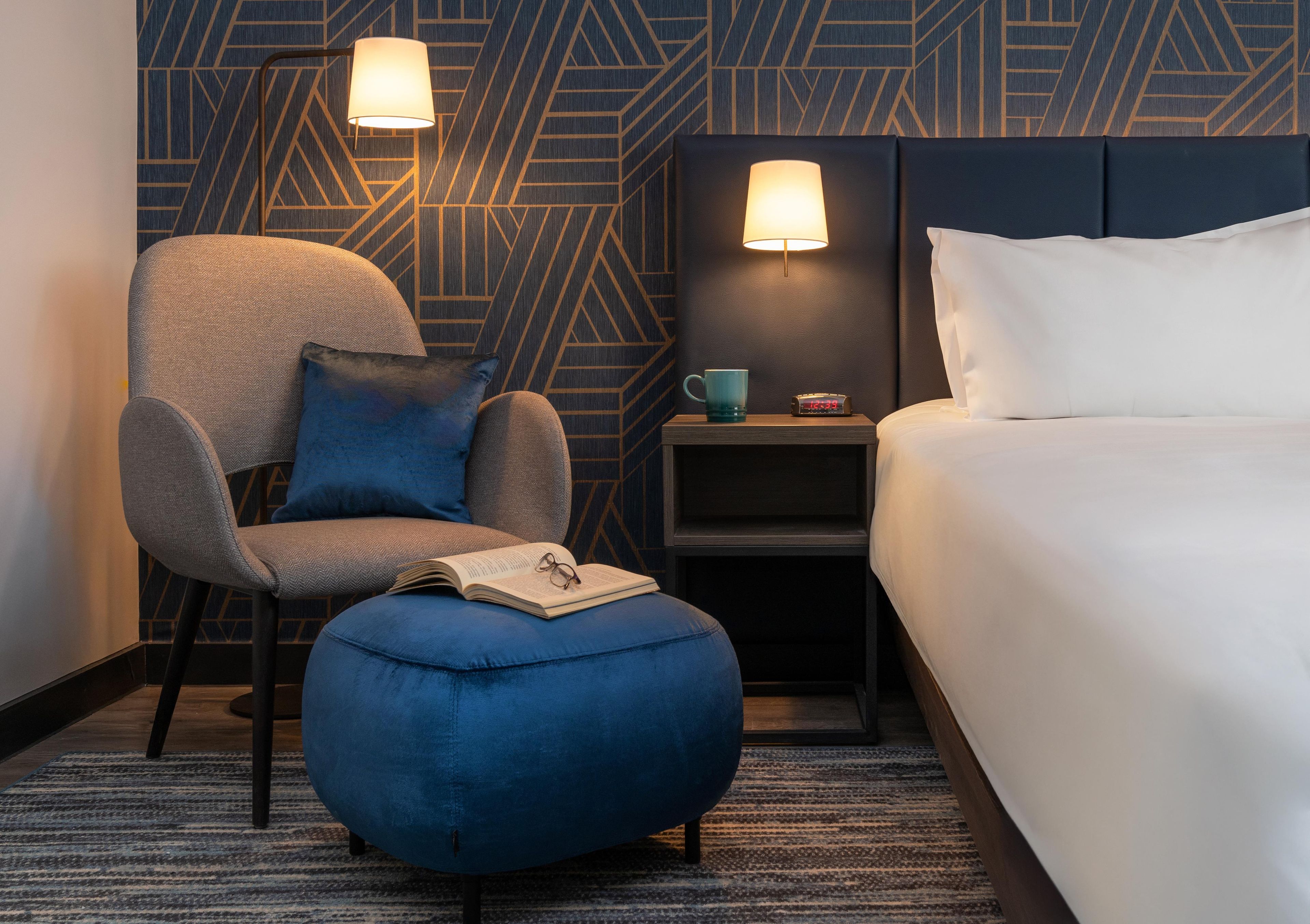 A hotel room with a bed, a chair, a side table with a lamp, a clock, and a book.