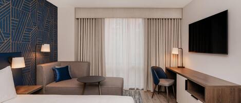 A hotel room with a bed, a sofa, a small table, a chair, a desk, a TV, and curtains.
