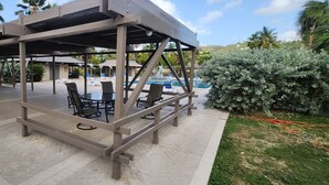 A covered outdoor seating area with tables and chairs, a pool in the background, and surrounding greenery. - Your tropical studio steps from the beach. (St Thomas)