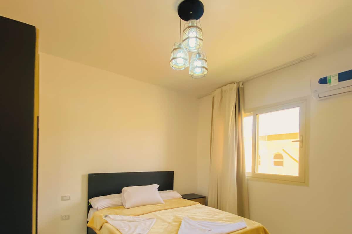 A bedroom with a bed, a bedside table, a ceiling light fixture, a window with curtains, and a door.