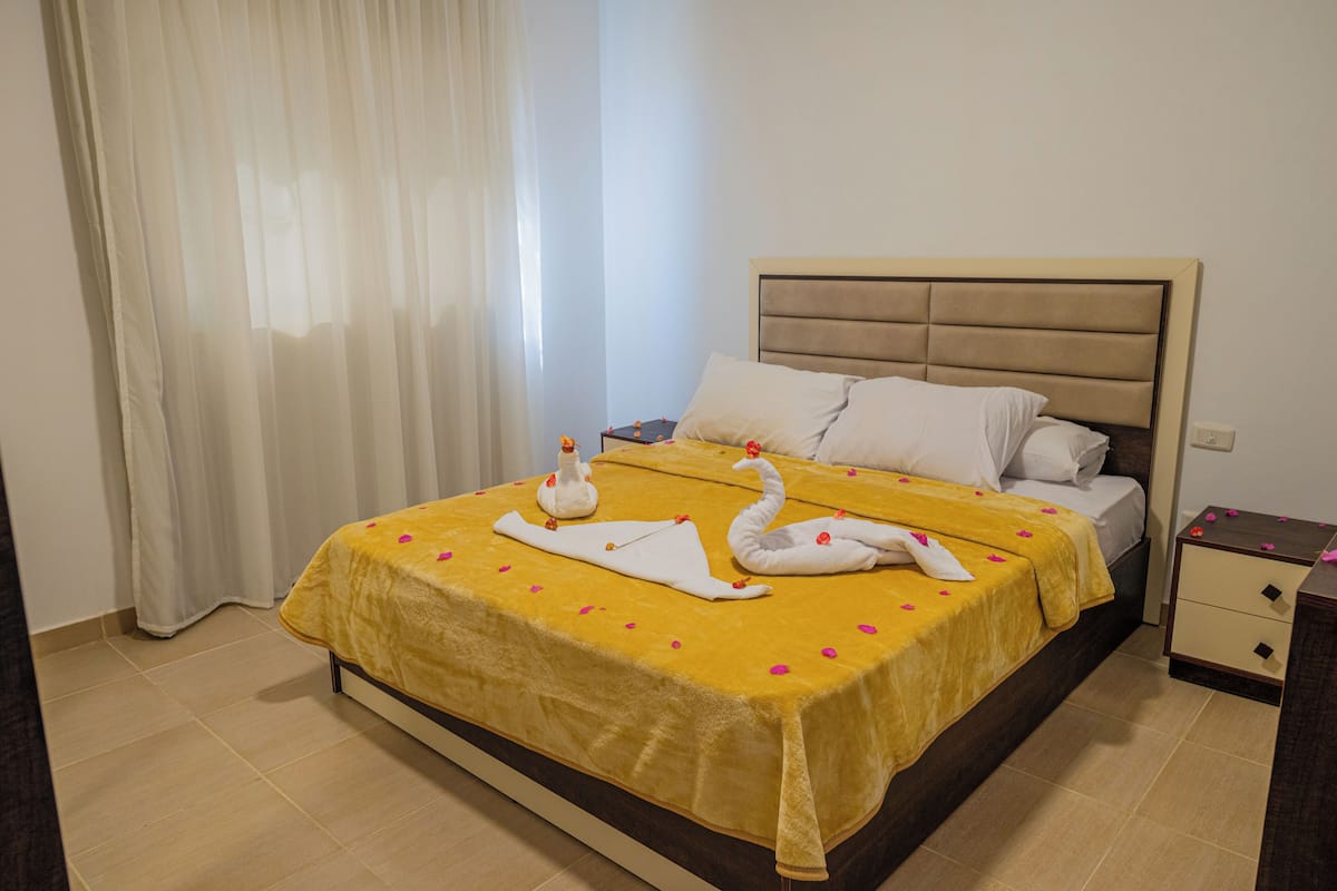 A neatly made bed with a yellow bedspread, two swan-shaped towels, and a small vase with flowers.