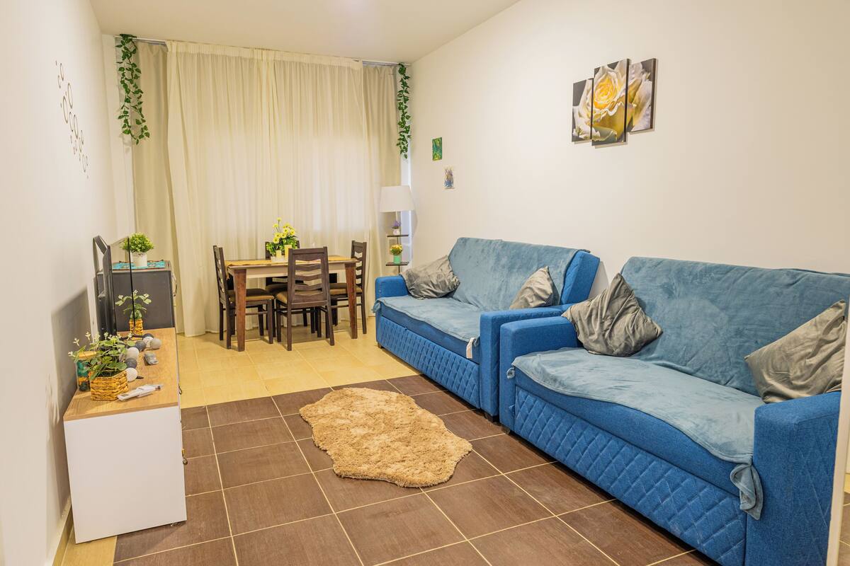 A living room with a blue sofa, a small white cabinet, a dining table with chairs, and a rug.