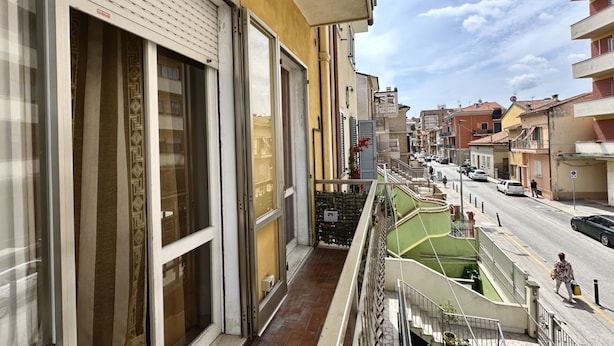 A balcony with a view of a street, a building with a patterned curtain, and a green area. - P032 - Porto Recanati, sky-land 100 meters from the sea (Porto Recanati)
