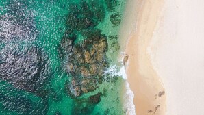 Vista aérea de una playa con agua turquesa cristalina, orilla arenosa y afloramientos rocosos.