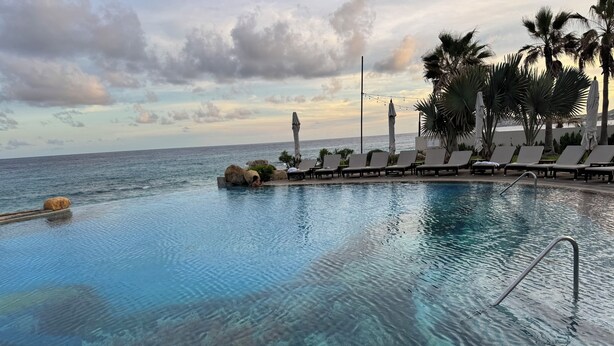 Una piscina cristalina con vista al océano, sillas de descanso y palmeras.