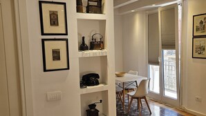 Um interior moderno com piso de madeira, uma parede branca com prateleiras exibindo objetos antigos e uma área de jantar com mesa e cadeiras.