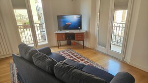 Sala de estar com um sofá cinzento, uma televisão sobre um suporte de madeira, e vista para a varanda através de amplas janelas.