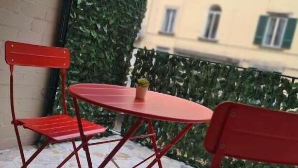 A small outdoor seating area with two red chairs and a table, a potted plant, and a view of a building with balconies. - Napul'è (Naples)