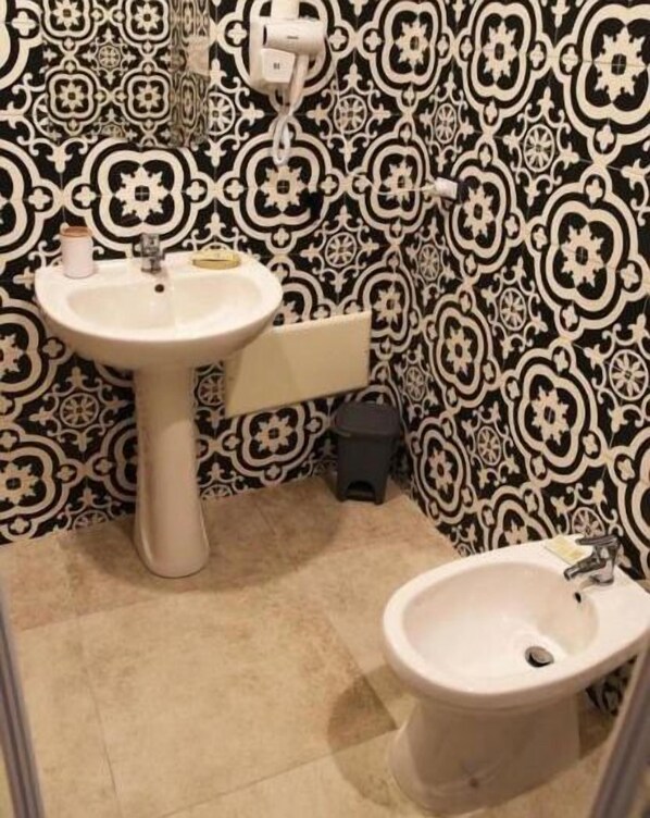 Um banheiro com pia de pedestal branca, bidê e vaso sanitário. As paredes possuem um papel de parede com padrão preto e branco.