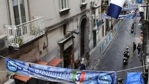 A narrow street with buildings on either side, a blue and white flag, and a banner with text. - Napul'è (Naples)