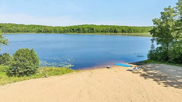 Blick auf einen See mit sandigem Ufer, einem kleinen Steg und einer Reihe von Liegestühlen.
