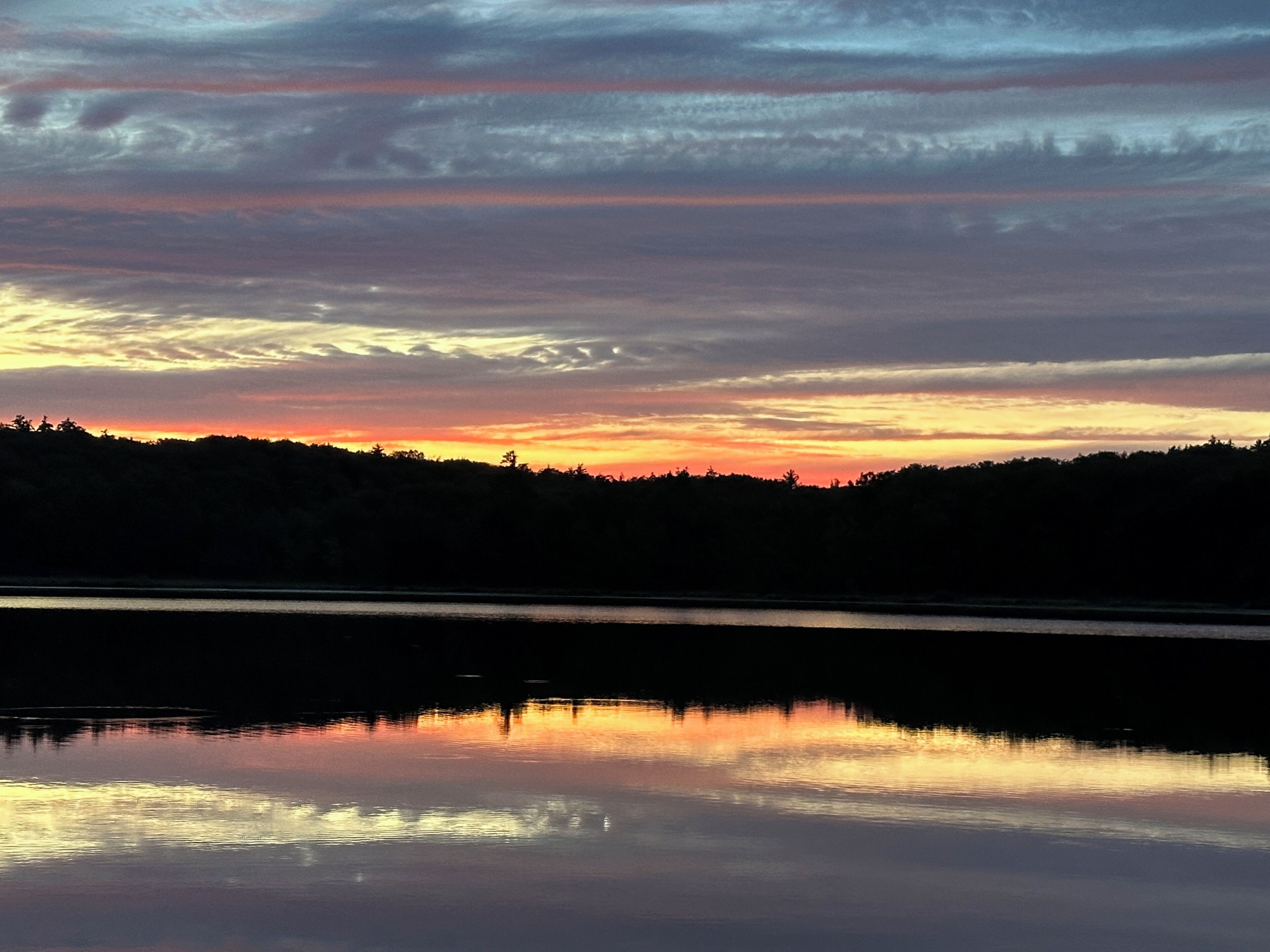 A calm lake reflecting the sunset sky with a silhouette of a forested hill in the background.