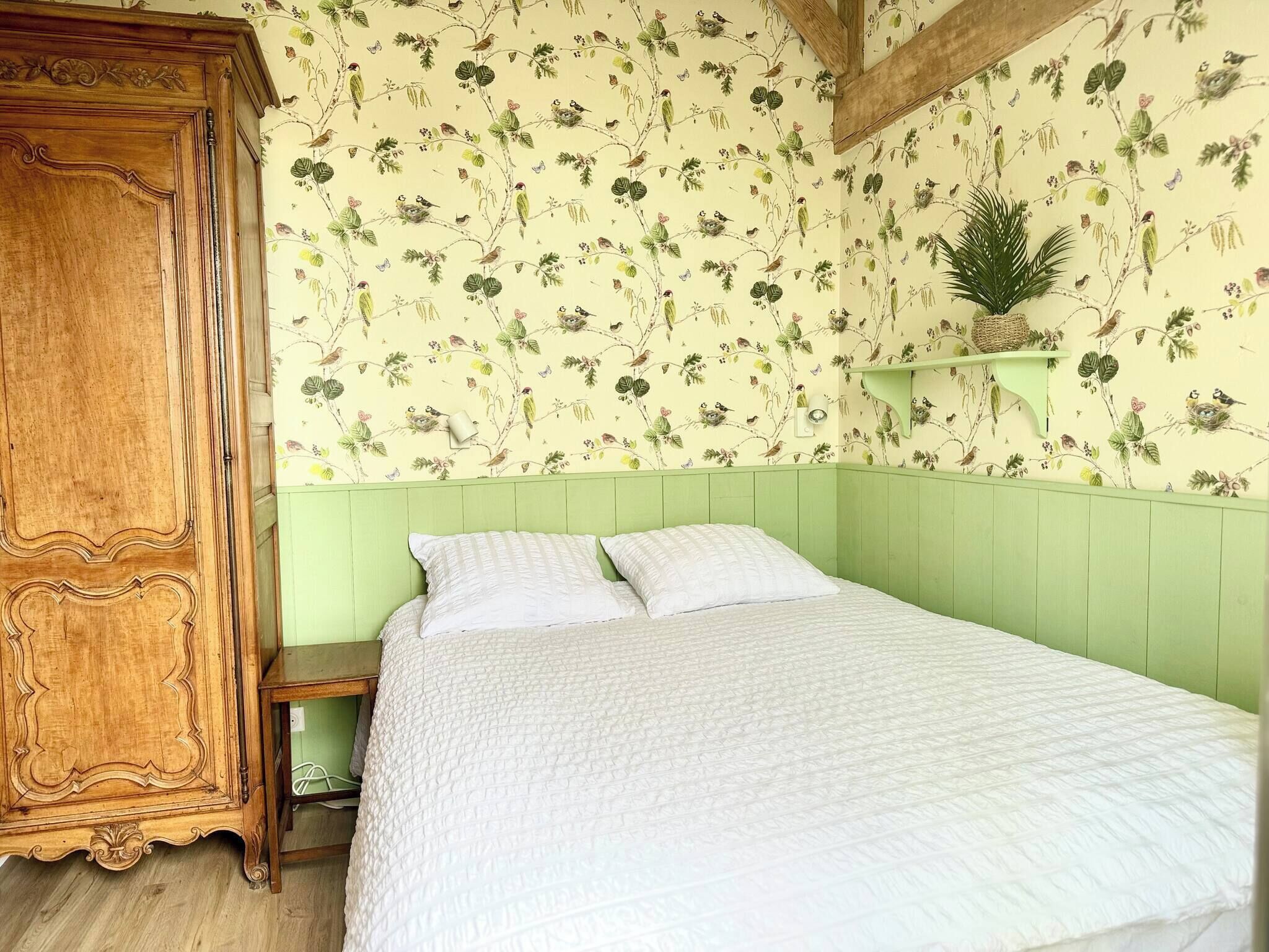 Une pièce comprenant un lit, une armoire en bois, une petite table et un mur recouvert d’un papier peint à motifs floraux.