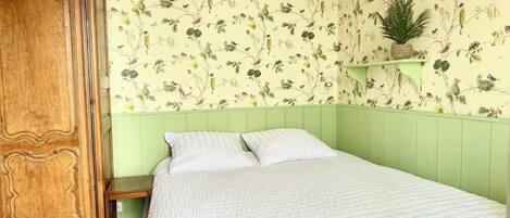 Une pièce comprenant un lit, une armoire en bois, une petite table et un mur recouvert d’un papier peint à motifs floraux.