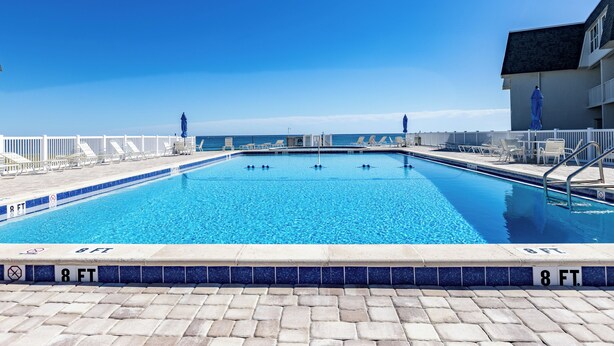 A rectangular swimming pool with a tiled edge, surrounded by a white fence, and a building with a dark roof in the background.