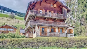 A multi-story wooden chalet with balconies, situated on a hillside with a view of a forested area.