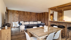 A modern living room with a wooden accent wall, a dark sofa with fluffy pillows, a glass dining table, and white chairs.