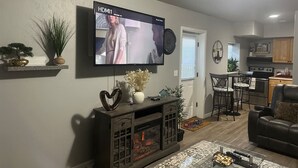 A modern living room with a flat-screen TV mounted on the wall, a fireplace, a dining area with a table and chairs, and a kitchen in the background.