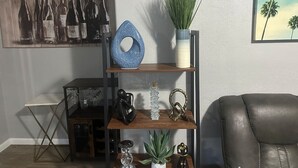 A living room with a wooden coffee table, a black leather armchair, a bookshelf with decorative items, and a painting on the wall.