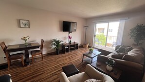 Sala de estar com piso de madeira, mesa de jantar com cadeiras, sofá, TV fixada na parede e varanda com vista para o exterior.
