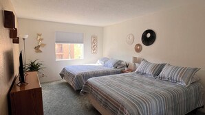 Quarto com duas camas, uma mesa de cabeceira de madeira, uma planta e uma janela com persianas.