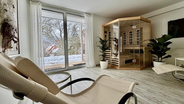 A modern living room with a large sliding glass door leading to a snowy outdoor area. There is a beige armchair, a small table, and a wooden cabinet with glass doors.