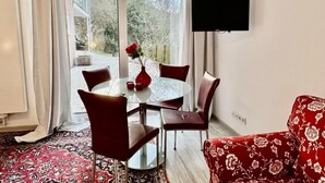 A dining area with a round table, chairs, and a vase with a red flower. A flat-screen TV is mounted on the wall.