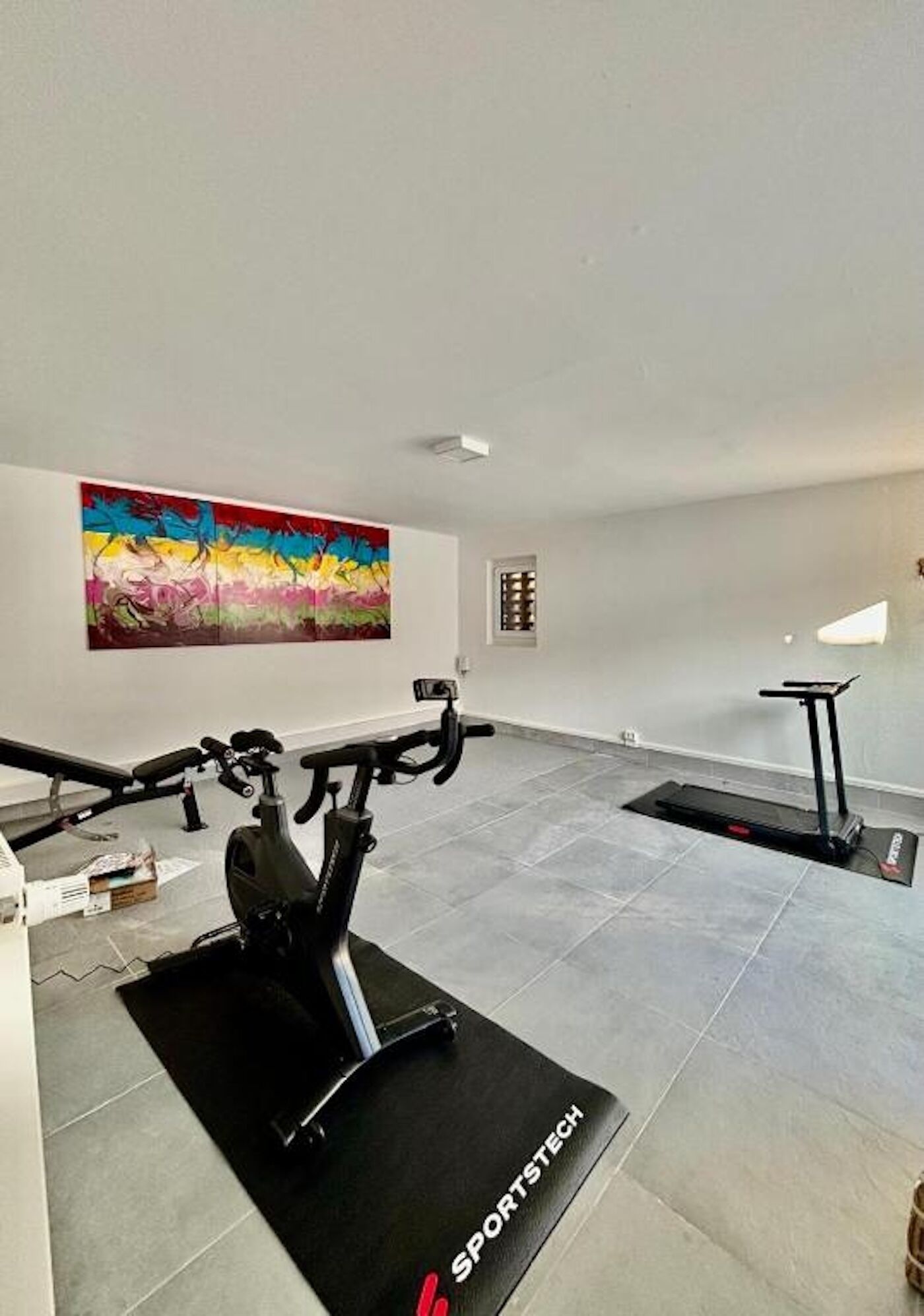 Un gimnasio en casa con equipo de ejercicio, un cuadro abstracto y colorido, y una ventana pequeña.