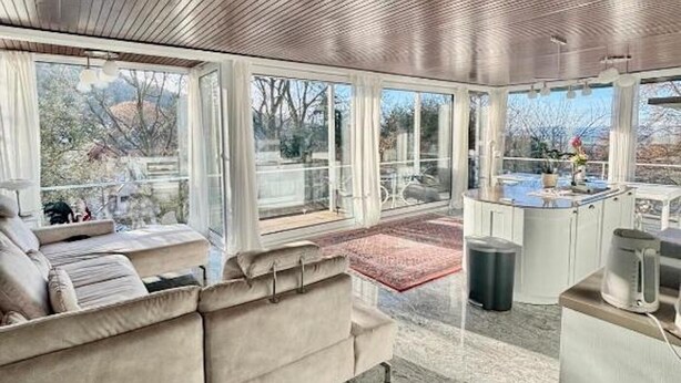 Un salon moderne avec un plafond en bois, un canapé beige, un coin cuisine et de grandes fenêtres donnant sur l’extérieur.