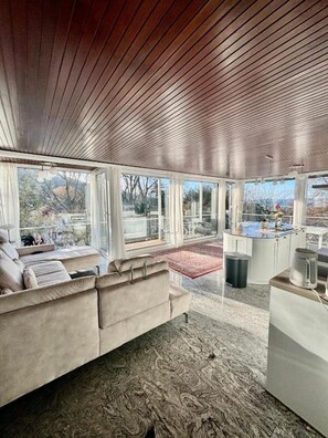 Una sala de estar moderna con techo de madera, un sofá color beige, una zona de cocina y ventanas amplias con vistas al exterior.