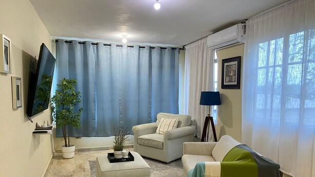 A living room with a sofa, armchair, and ottoman, a flat-screen TV, a lamp, and a potted plant.