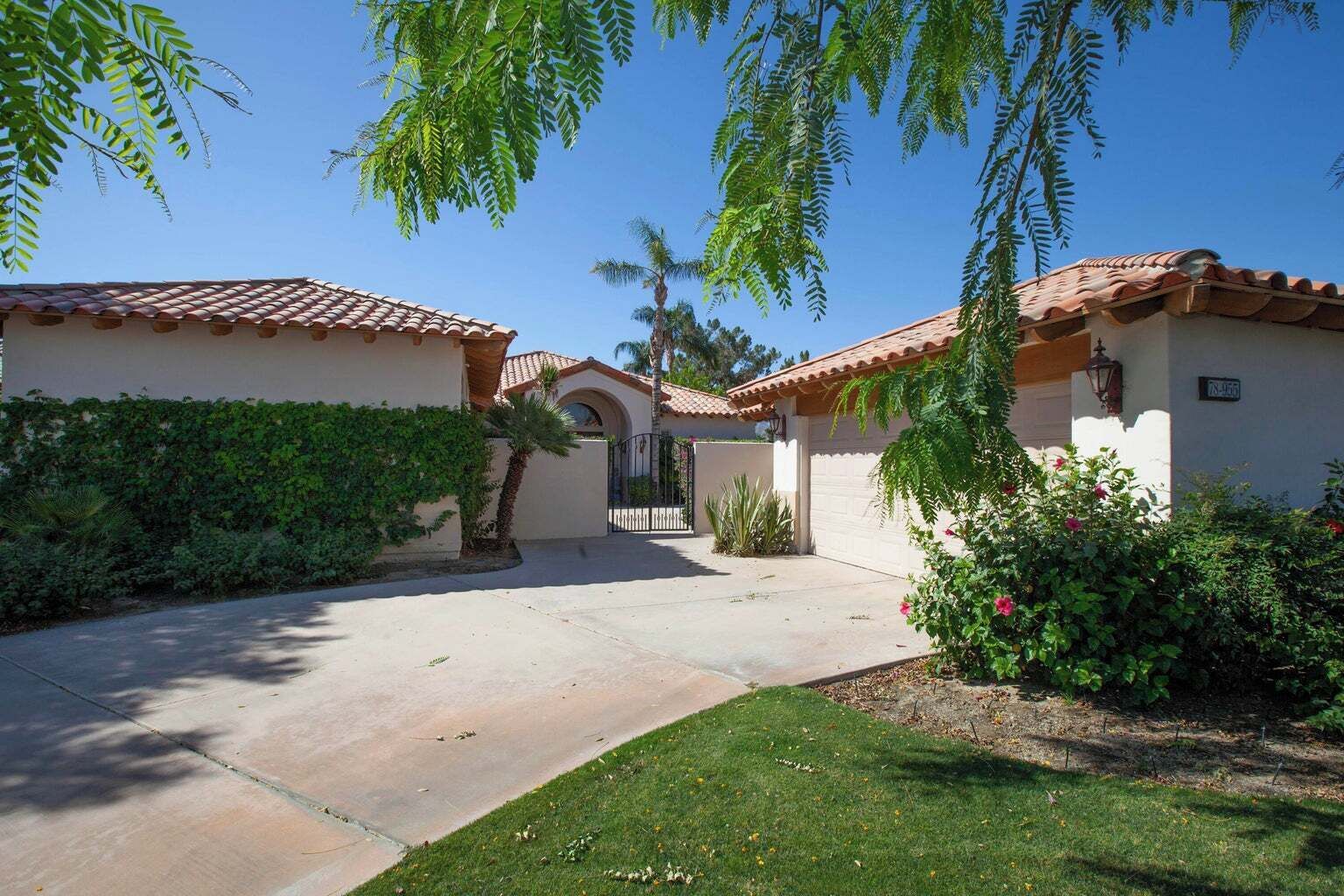 A residential area with a paved driveway leading to a gated entrance, surrounded by lush greenery and palm trees.