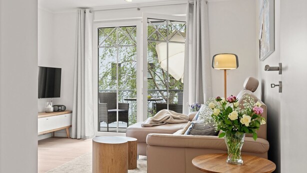 A modern living room with a sofa, a coffee table, and a TV. There is a balcony with a view of trees and a chair. - Ostseeliebe Wg. 6 /1. Etage - Neue, Hochwertig Eingerichtete Wohnung in Strandnähe (Zingst)