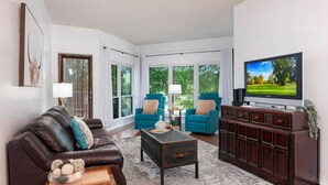 A living room with a brown leather sofa, two teal armchairs, a coffee table, and a TV on a wooden cabinet.