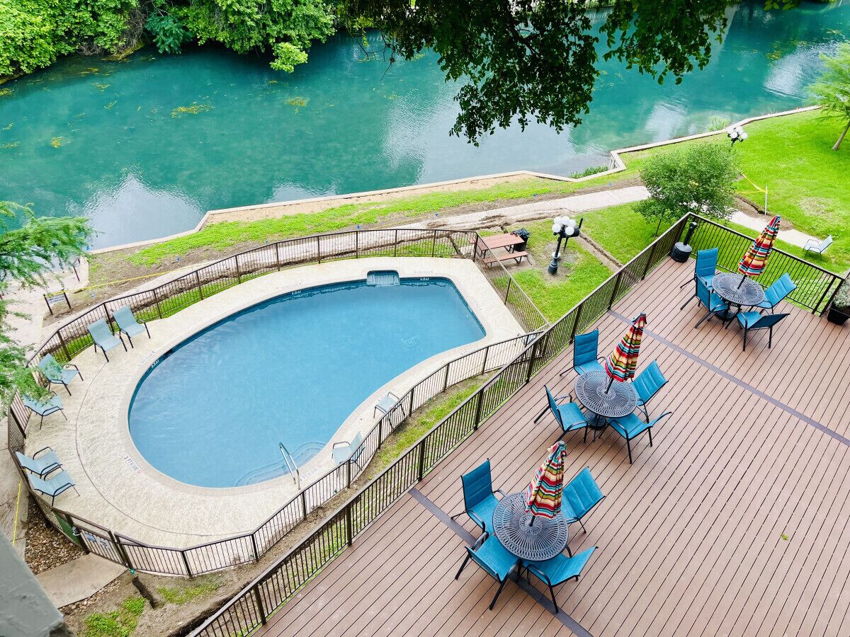 A swimming pool with a curved edge, surrounded by a deck with chairs and tables, and a view of a river.
