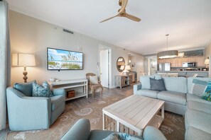 A spacious living room with a flat-screen TV, a ceiling fan, and a kitchen in the background. - Grand Pointe 205 | Beachfront, Pools, Saunas, Hot Tub | My Beach Getaways (Orange Beach)