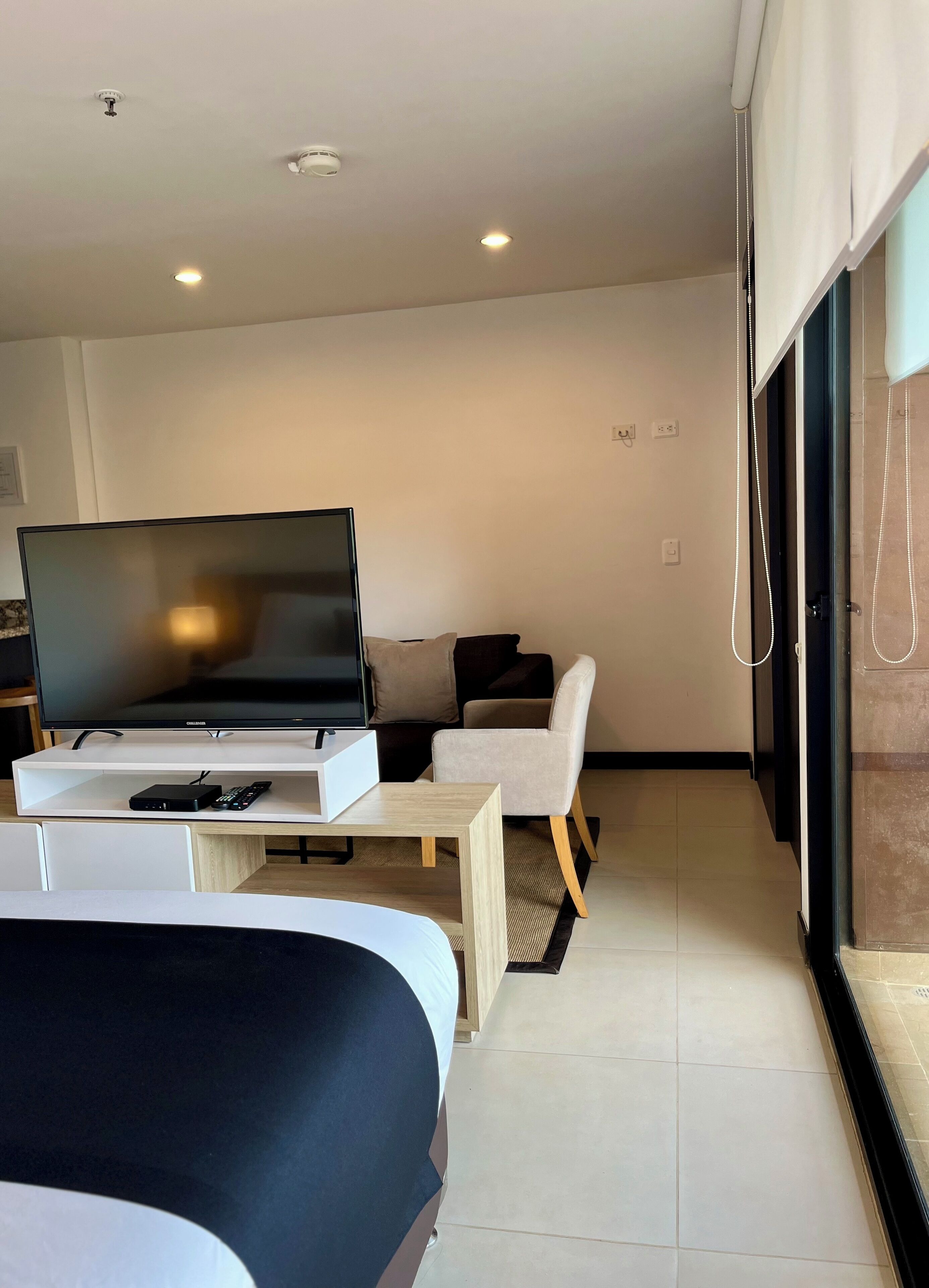 A modern hotel room with a flat-screen TV, a bed, a small table, a chair, and a large window.