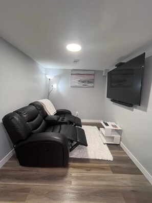 A modern living room with a black leather sofa, a flat-screen TV mounted on the wall, and a small white side table with a remote control. - Adorable Private Suite in Seton SE, Calgary
 (Calgary)