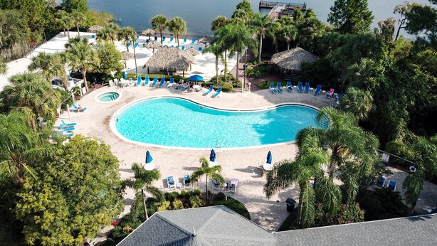Un resort con una amplia piscina rodeada de sillones de descanso y sombrillas, con vistas a un lago y árboles frondosos.