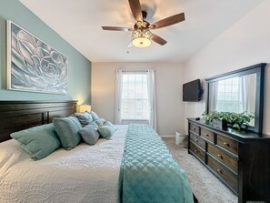 Ein Schlafzimmer mit einem Bett, einem Schminktisch, einem Deckenventilator, einem Fernseher und einem Fenster mit Jalousien.