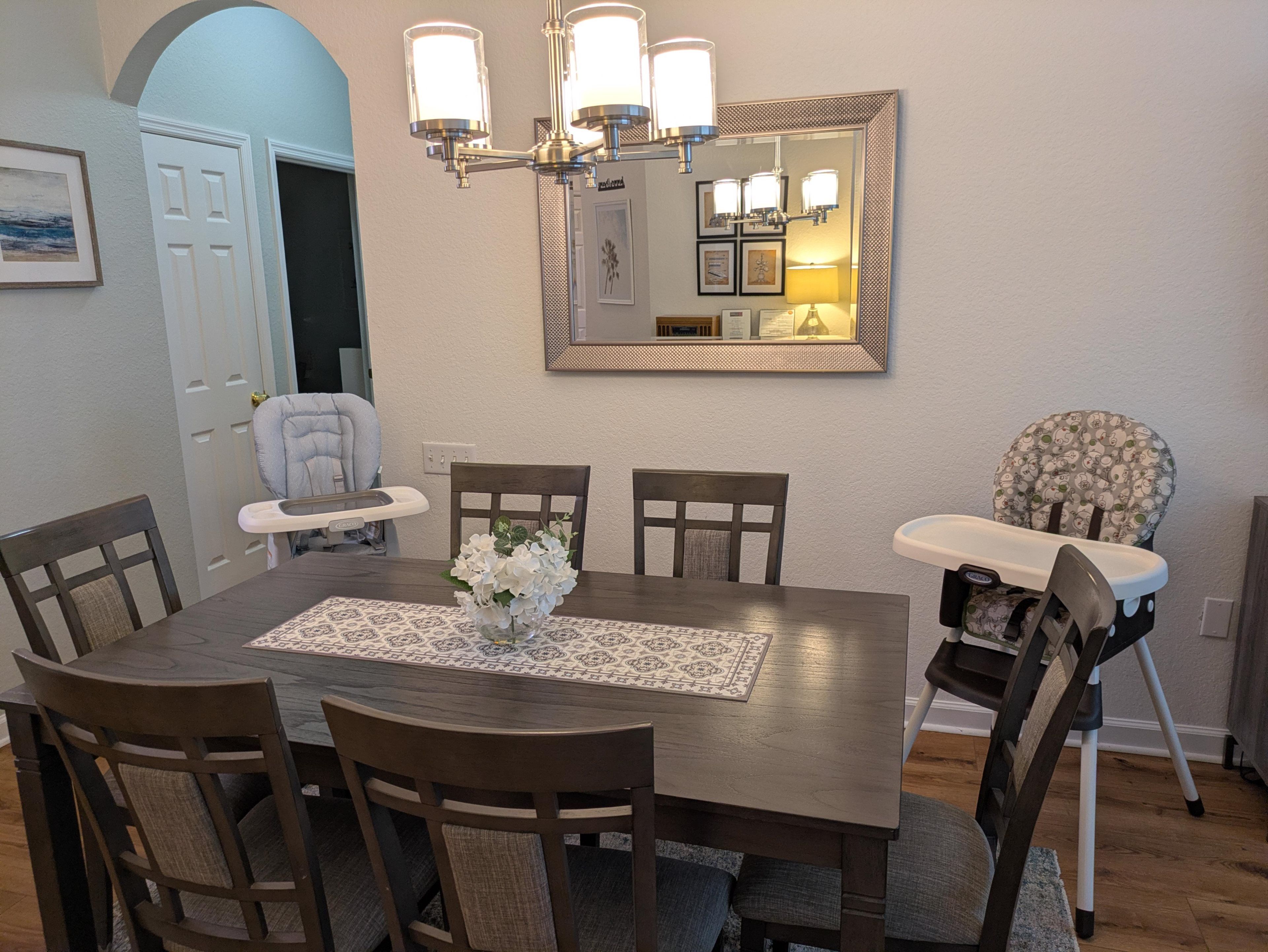 A dining room with a wooden table, chairs, a vase of flowers, a mirror, and a baby high chair.