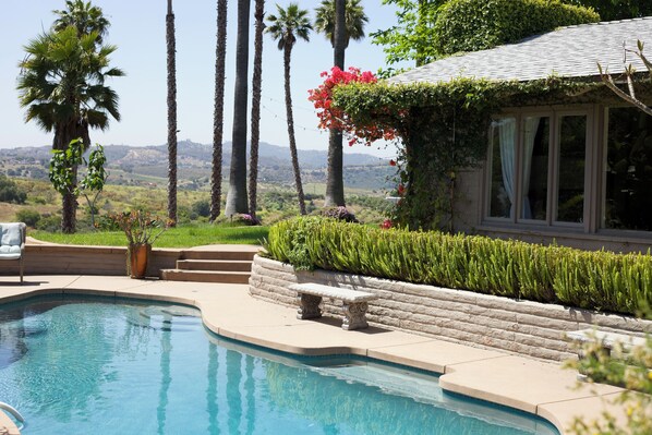 A swimming pool with a stone bench, surrounded by a landscaped garden with palm trees and a view of distant hills.